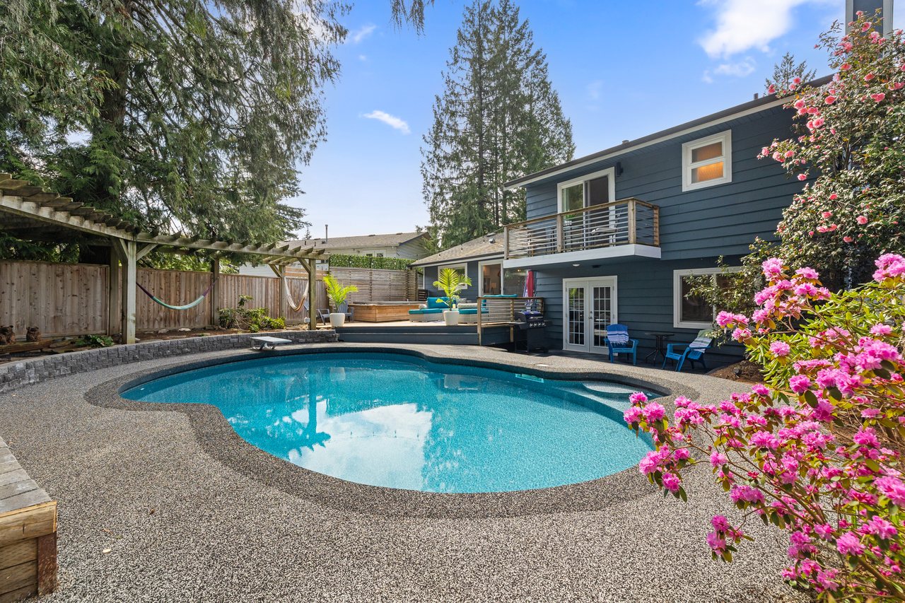 Backyard of a blue two-storey house with a kidney-shaped pool, a pergola, tall evergreens and pink rhododendrons in bloom.